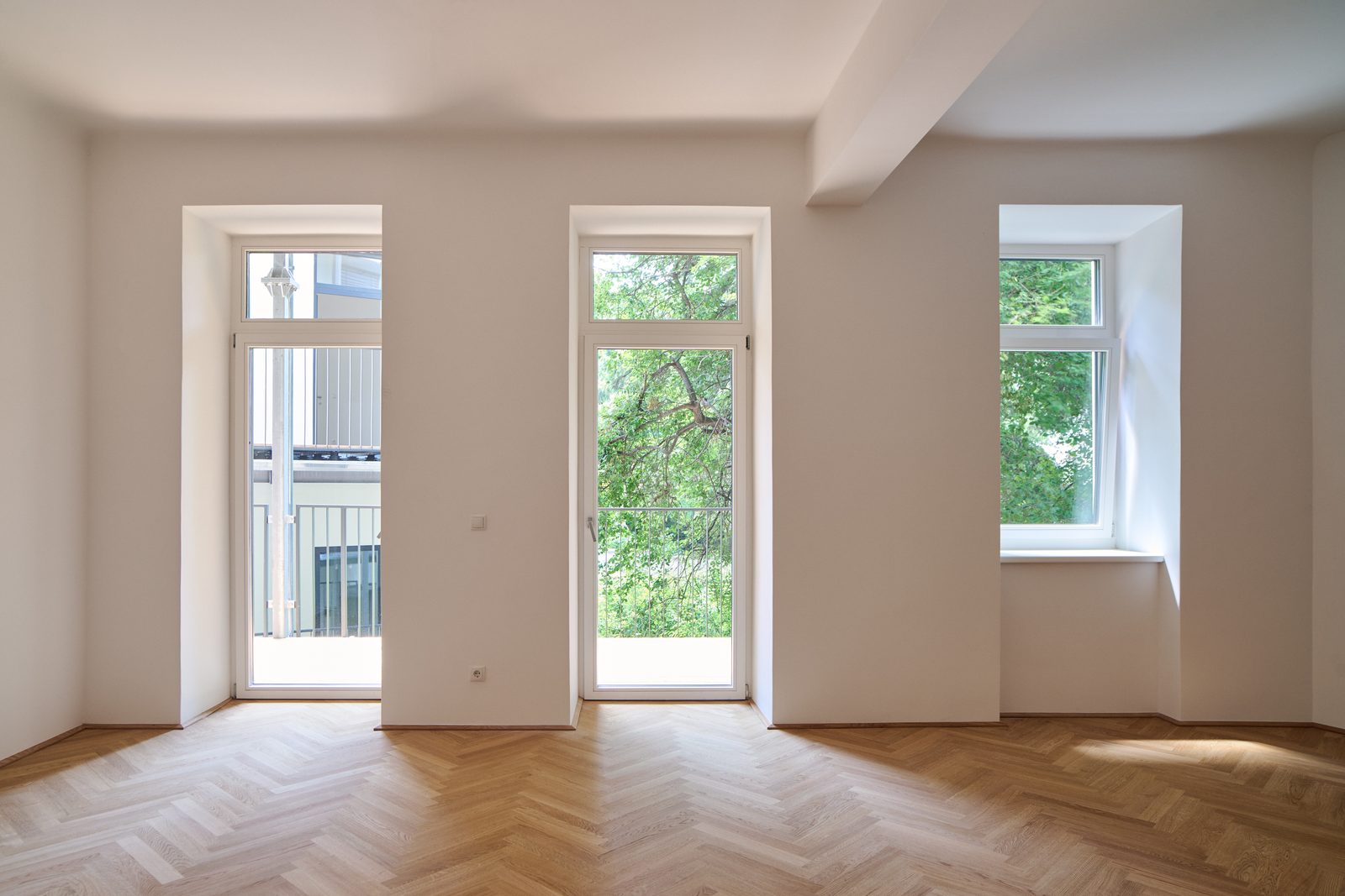 Wohnraum mit Fischgrätparkett, zwei Balkontüren und Fenster ins Grüne